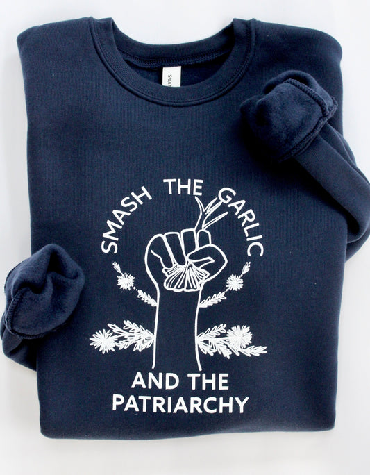A folded navy blue crewneck sweatshirt reads "Smash the Garlic and the Patriarchy" in white lettering with an illustration of a hand holding garlic