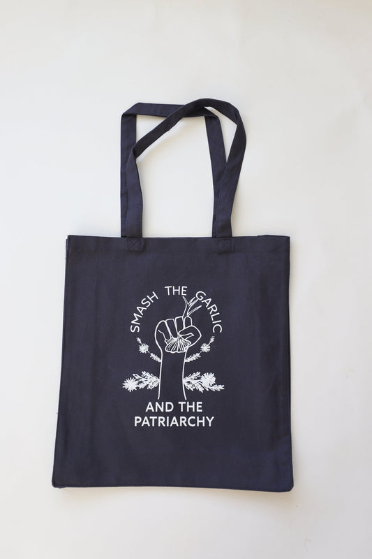 A navy blue canvas tote with white lettering reads "Smash the Garlic and the Patriarchy" with a garlic illustration 