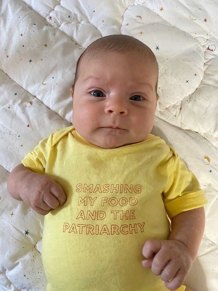 A baby lays on a quilt wearing a yellow onesie that reads "Smashing my Food and the Patriarchy"