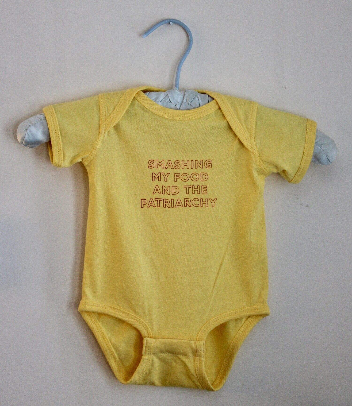 A yellow baby onesie that reads "Smashing my Food and the Patriarchy" hangs on a hanger