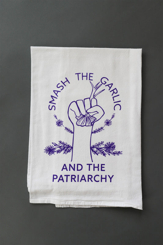 A white tea towel with purple lettering reads "Smash the Garlic and the Patriarchy" with an illustration of a hand holding garlic