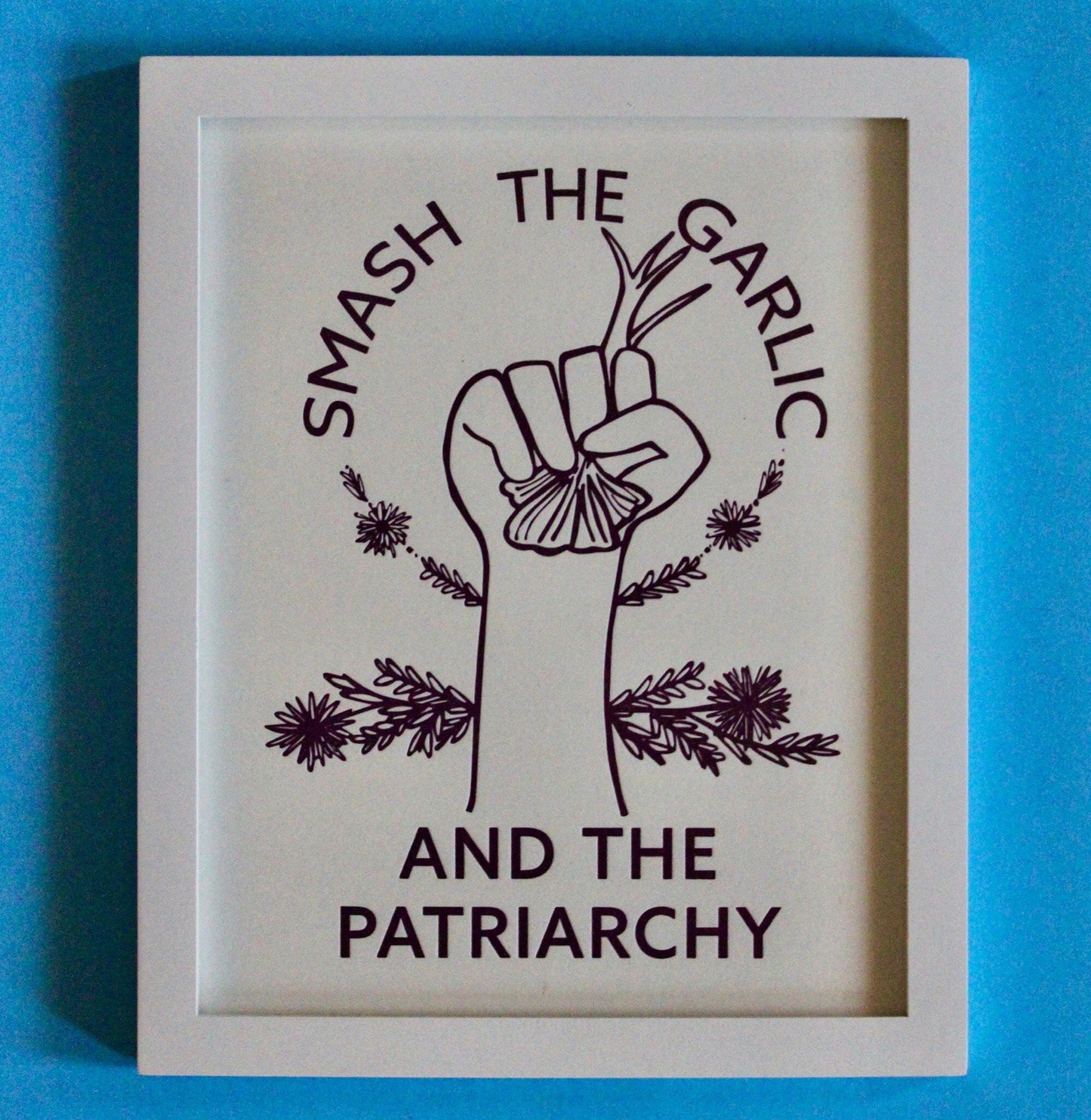 An art print in a frame reads "Smash the Garlic and the Patriarchy" with an illustration of a hand holding garlic 