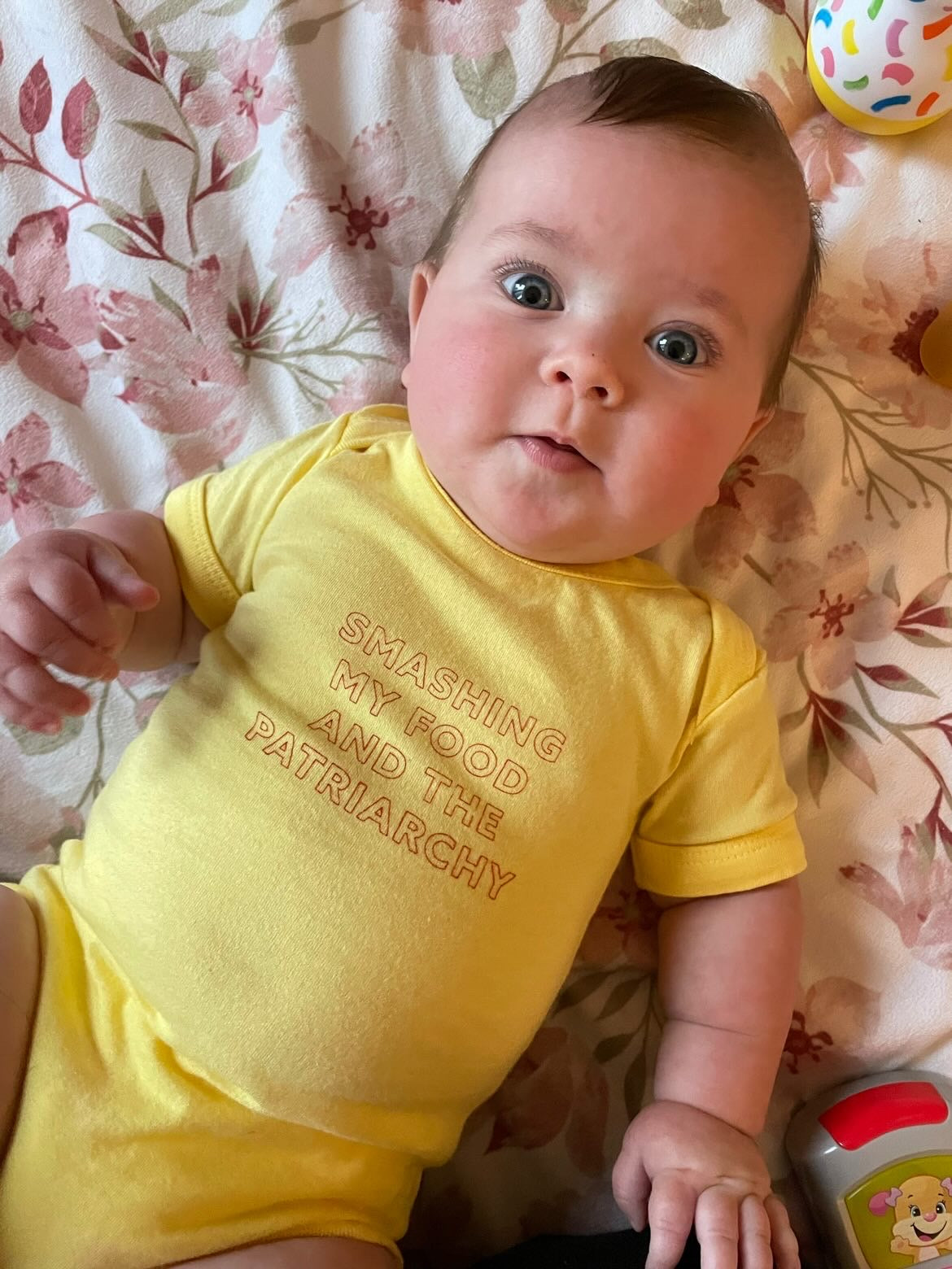 A baby wears a yellow onesie that reads "Smashing my Food and the Patriarchy"