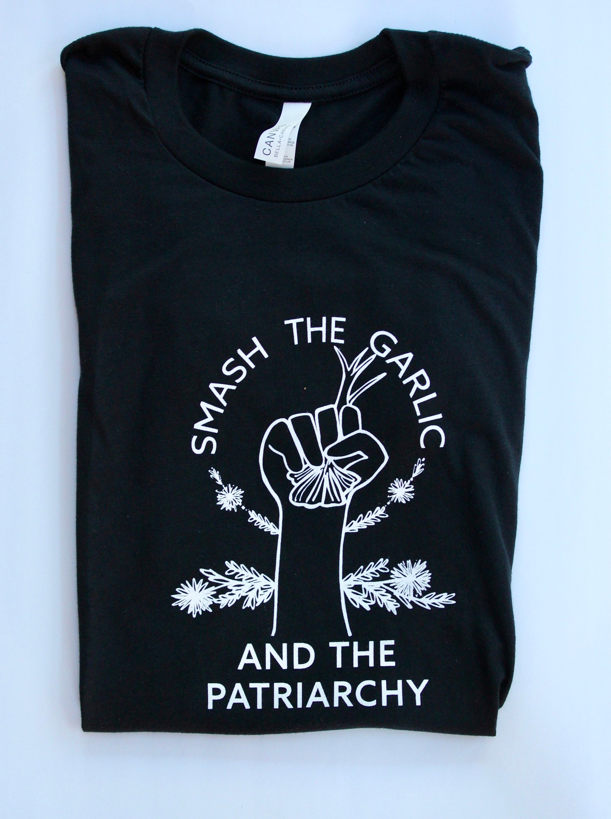 A folded black tee with white lettering reads "Smash the Garlic and the Patriarchy" with a garlic illustration 