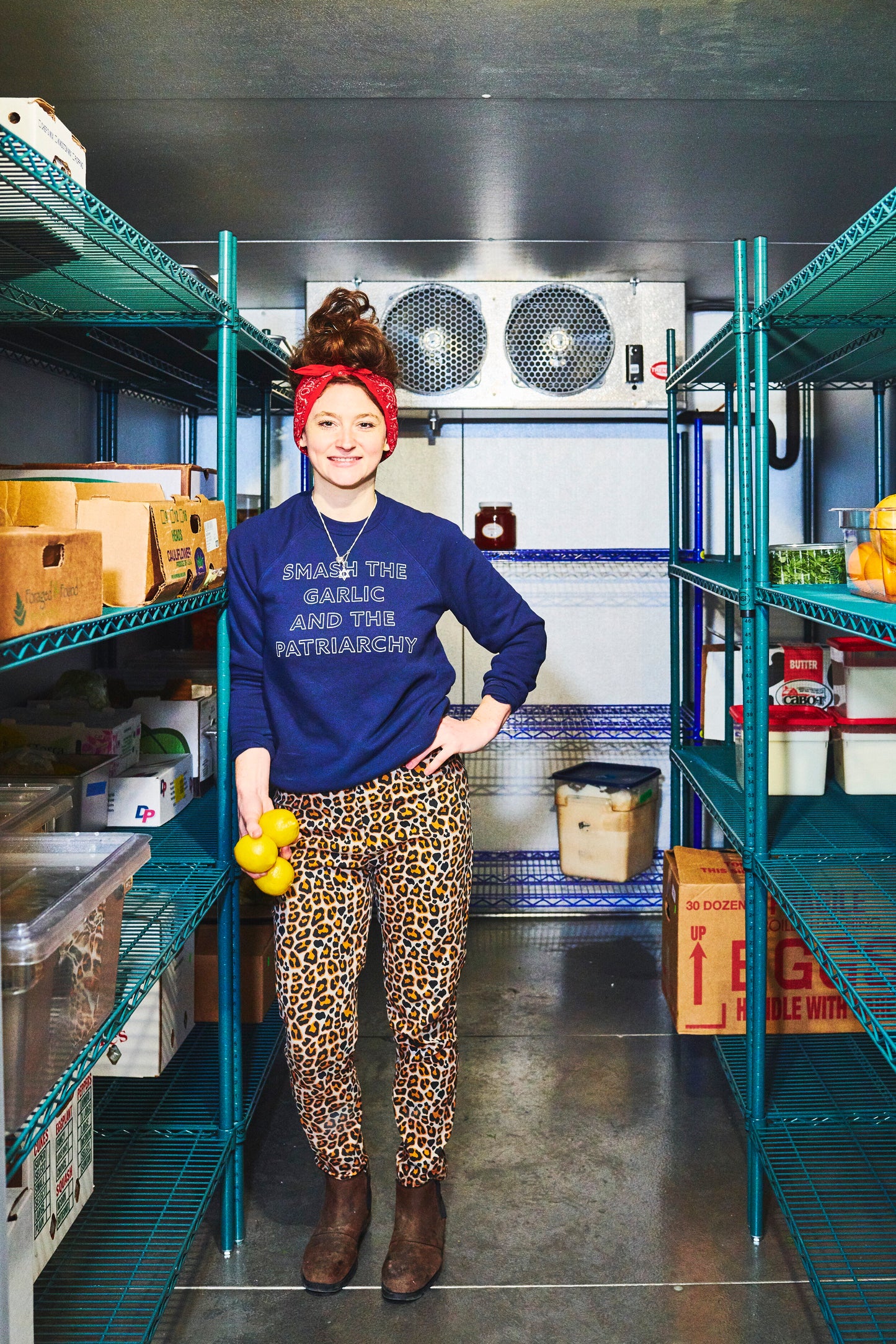 A woman wearing a navy blue "Smash the Garlic and the Patriarchy" stands in a walk-in refrigerator 