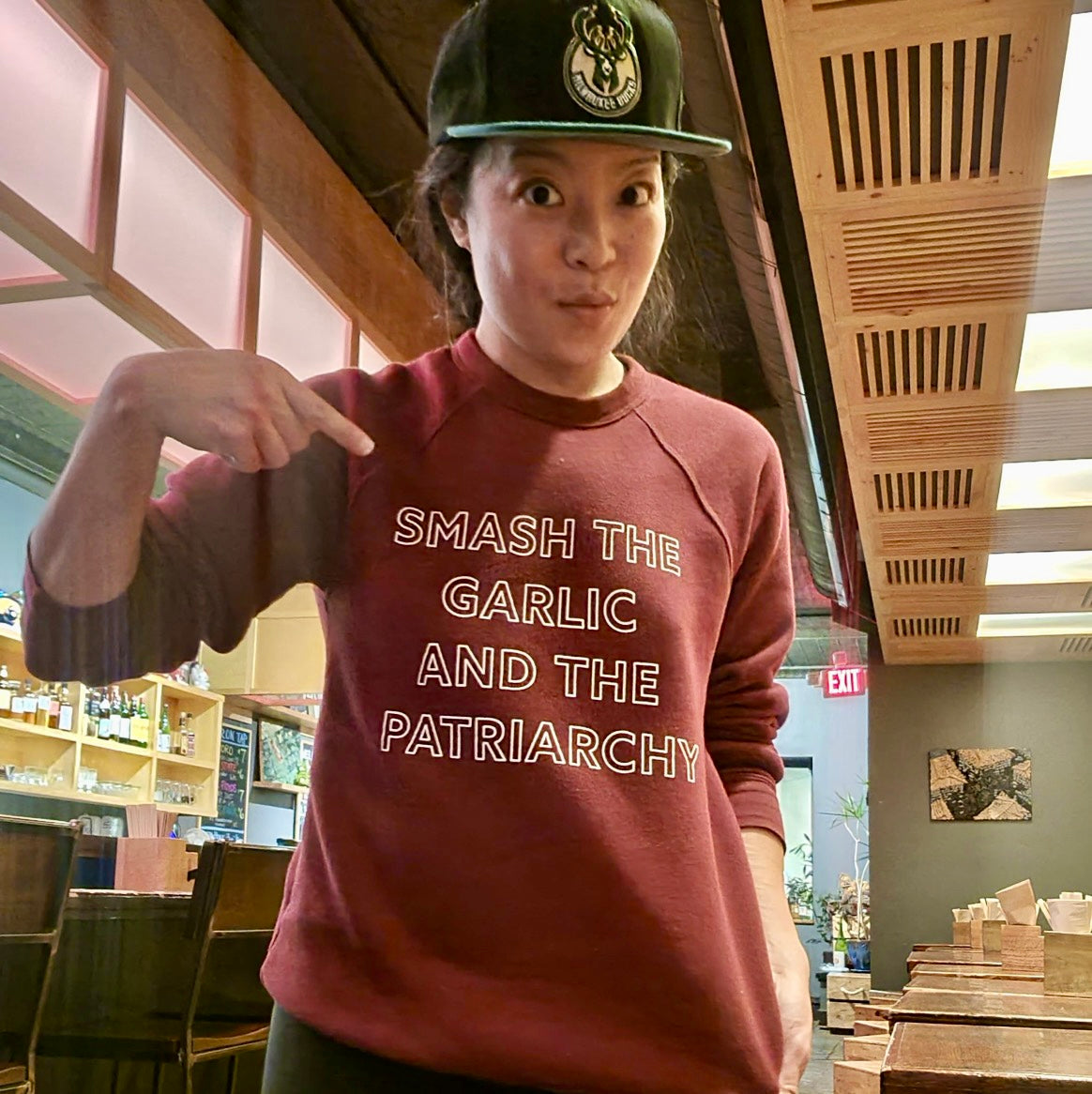 A woman wears a maroon crewneck that reads "Smash the Garlic and the Patriarchy" in white block letters