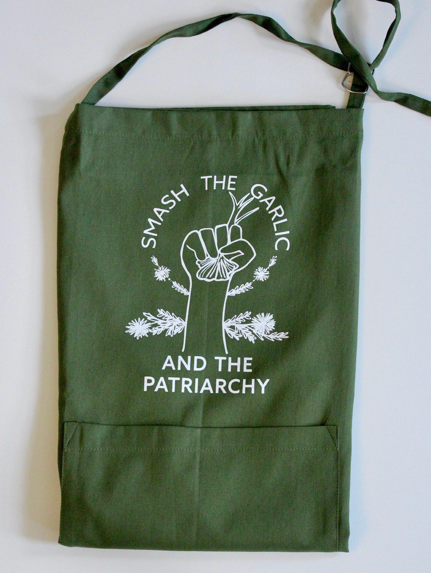A folded dark green apron with the words "Smash the Garlic and the Patriarchy" and an illustration of a hand holding garlic 