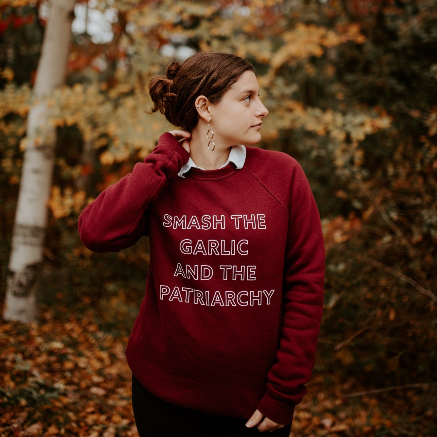 A woman wearing a maroon "Smash the Garlic and the Patriarchy" stands in a colorful forest