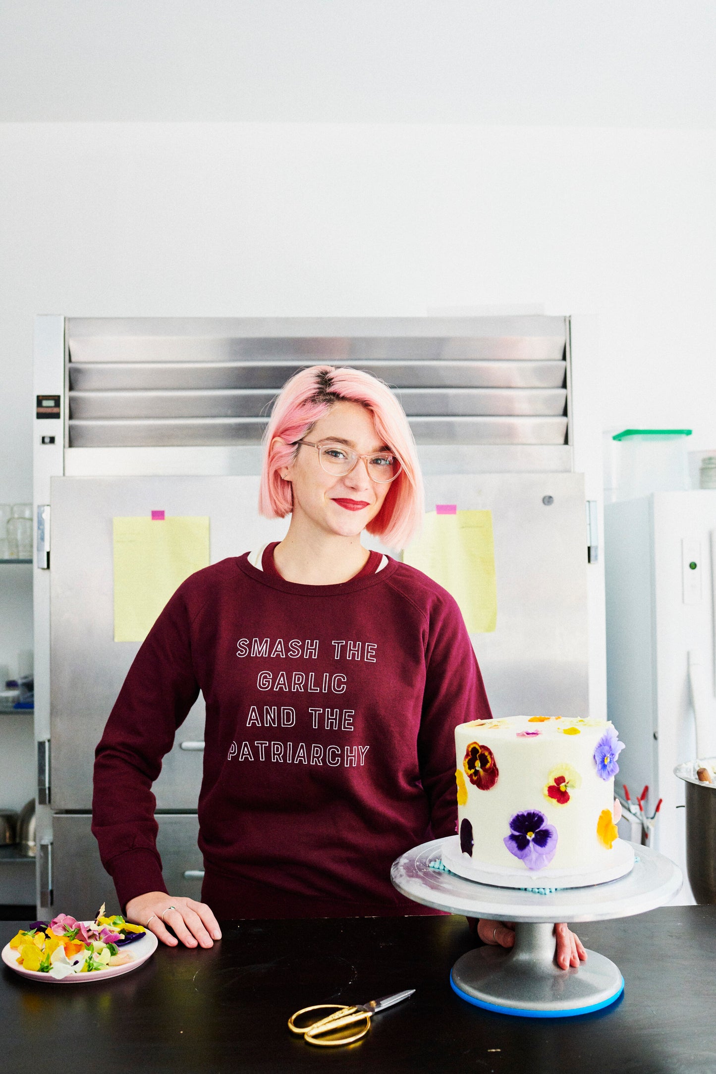 A woman wearing a maroon "Smash the Garlic and the Patriarchy" crewneck stands with a cake
