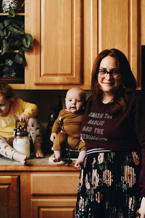 A woman wearing a maroon "Smash the Garlic and the Patriarchy" sweatshirt stands with a baby and toddler