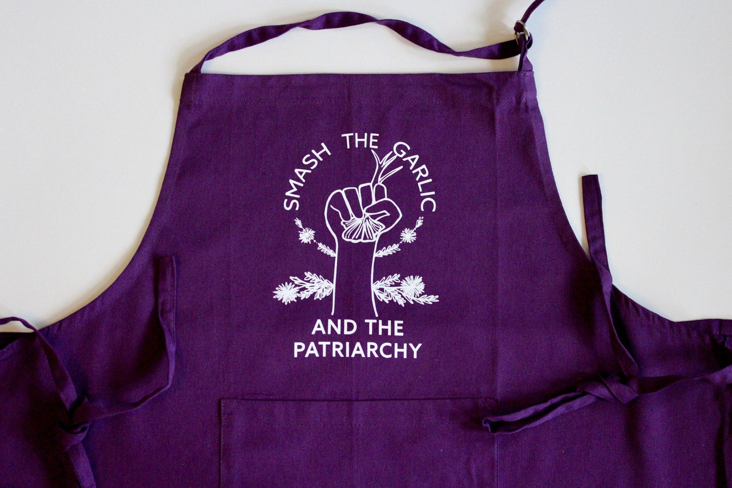 A dark purple plus size apron that reads "Smash the Garlic and the Patriarchy" in white lettering with an illustration of a hand holding garlic