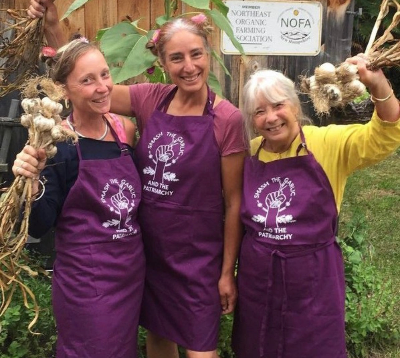 Three women wear purple "Smash the Garlic and the Patriarchy" aprons and hold bunches of garlic
